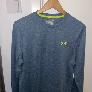 Blå långärmad träningströja Under Armour - Snygg blå långärmad träningströja från Under Armour med gul logga på bröstet. Perfekt för gymmet eller löprundan. Tillverkad i ett lätt och snabbtorkande syntetmaterial som andas. Rund halsringning och normal passform.