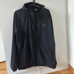 Svart vindjacka från Under Armour - Svart vindjacka från Under Armour med huva och dragkedja framtill. Jackan har en clean look med diskret logga på bröstet, snörning i huvan och två sidofickor. Perfekt för blåsiga dagar och enkel att matcha med allt. Varan har två små defekter skicka dm vid intresse. Pris kan diskuteras.