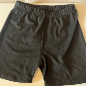 Svarta stretchiga shorts i polyester - Svarta shorts med elastisk midja, tillverkade i mjukt och stretchigt material (92% polyester, 8% spandex). De har en enkel och clean look, perfekt för dig som gillar basic och bekvämt. Passformen är normal och tyget är lätt och följsamt.