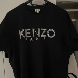 Svart Kenzo t-shirt med logga - Svart t-shirt från Kenzo med stor vit logga 'KENZO PARIS' på bröstet. Klassisk rund hals och korta ärmar. Snygg och stilren design som passar perfekt till jeans eller shorts. Tillverkad i mjuk bomull för skön känsla hela dagen.