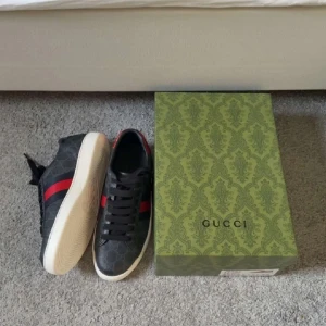 Gucci sneakers med röd/blå rand - Snygga svarta Gucci sneakers med klassisk röd och blå rand på sidan och diskret GG-mönster över hela skon. Yttermaterialet är canvas med detaljer i skinn och textil, vit platt sula och svarta skosnören. Bakpartiet har metallicröd och blå detalj. 