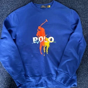 Blå sweatshirt från Polo Ralph Lauren - Blå sweatshirt från Polo Ralph Lauren med färgglatt tryck framtill i orange, gul och vit. Klassisk rund halsringning och långa ärmar. Mjuk och skön bomullskvalitet, perfekt för en chill och stilren look.