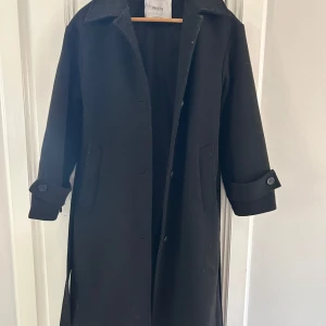 Svart kappa från Bershka med bälte - Säljer en svart lång kappa från Bershka i klassisk modell med bälte i midjan och knappar vid ärmsluten. Jackan har två fickor framtill och är perfekt för kyligare dagar. Enkel och stilren design som passar till många outfits.