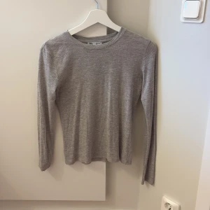 Grå/beige långärmad tröja från Zara - Enkel och stilren grå långärmad tröja från Zara i storlek S. Tröjan har rund halsringning och är i ett mjukt, tunt bomullsmaterial. Perfekt att styla med jeans eller kjol för en clean look.