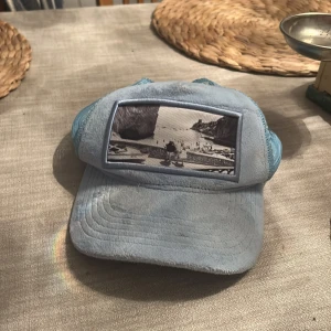 Ljusblå  Emilio Pendenza keps - Snygg ljusblå keps från Emilio Pendenza med mesh på sidorna och justerbar snapback. Framtill finns ett svartvitt foto-tryck som ger en cool vibe. Perfekt för dig som vill sticka ut med en unik accessoar. Kepsen är tillverkad i syntetmaterial och har en klassisk böjd skärm. Inga fel