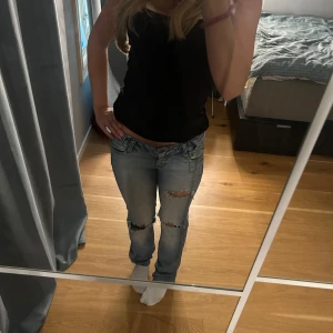 Lågmidjade slitna jeans - Säljer ett par ljusblå jeans med slitningar över knäna och klassiska femficksdetaljer. Jeansen har låg midja och bootcut. Har en liten defekt och ett lagat hål som inte märks av.