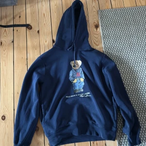 Blå Ralph Lauren hoodie - Mörkblå hoodie från Ralph Lauren med ikoniska Polo Bear-trycket framtill. Storlek S. Liten defekt sista bilden. Sitter perfekt på mig som är 172 och väger 62