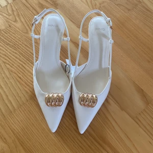 Vita pumps med gulddetalj från MNG - Snygga vita pumps från MNG med spetsig tå och smal klack. Skorna har en elegant guldfärgad detalj framtill och tunna remmar runt hälen. Perfekta för dig som vill sticka ut med en stilren och trendig look. Helt nya (nypris 299kr)