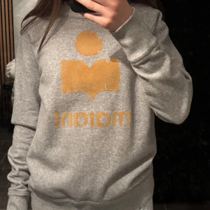 Grå sweatshirt Isabel Marant - Snygg grå sweatshirt från Isabel Marant Étoile med stort gult logotryck framtill. Tröjan har rund hals, långa ärmar och en klassisk passform. Perfekt för en chill och trendig look. Det är meningen att loggan/märket ska vara spruket.