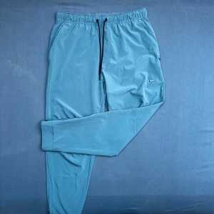 Blå Nike tech/tränings byxor med snörning - Snygga ljusblå tech/träningsbyxor från Nike med svart snörning i midjan och broderad Swoosh-logga på benet. Byxorna har elastisk midja, smal passform och dragkedja vid benslut. Perfekta för chill eller träning. Ett litet hål finns på baksidan men syns ej när byxorna bärs, om man kollar noga kan man nog hitta en liten fläck vid hålet, utöver det är de i otroligt bra skick