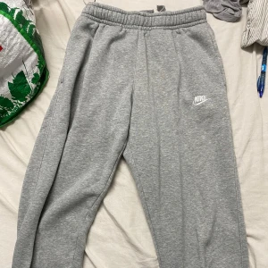 Grå mjukisbyxor från Nike - Gråa mjukisbyxor från Nike med elastisk midja och dragsko. Byxorna har en klassisk passform, sidofickor och en bakficka med knapp. Mjukt och skönt material, perfekt för chill eller träning. Nike-logga broderad på ena benet.