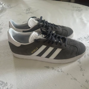 Adidas Gazelle  - Snygga Adidas Gazelle sneakers i mörkgrå mocka med vita detaljer och klassiska tre ränder på sidorna. Skorna har svart snörning, vit plös med Adidas-logga och vit sula. Perfekta för dig som gillar retrostil och streetwear. Bra skick, använda sparsamt, strl 43 1/3, finns ingen box