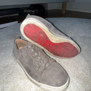 Grå mockasneakers med röd sula - Snygga sneakers i grå mocka med klassisk snörning och vit sula. Insidan är i beige skinn och yttersulan har en cool röd detalj undertill. Skorna har en rund tå och stilren design som passar till det mesta.