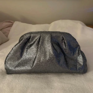 Glittrig silver kuvertväska - Snygg kuvertväska i glittrigt silver med mjuk form och lätt rynkade detaljer upptill. Perfekt när du vill addera lite extra bling till din outfit. Väskan är stilren och enkel att matcha med olika looks.