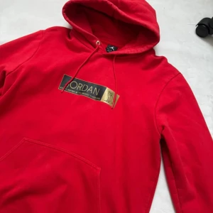 Röd Jordan OVO hoodie - Riktigt snygg röd hoodie från Jordan med stor huva och snörning. Framtill finns en svart och guldig logga med text och uggletryck. Klassisk känguruficka och mjukt bomullsmaterial gör den både bekväm och stilren. Perfekt för dig som gillar streetwear vibes.