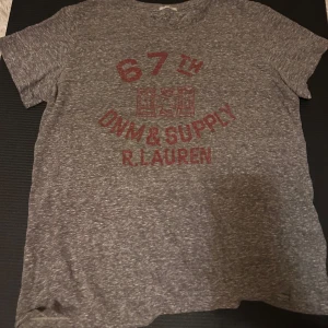 Grå t-shirt från Ralph Lauren - Snygg gråmelerad t-shirt från Ralph Lauren Denim & Supply med röd tryckt text och grafiskt motiv på bröstet. Modellen är RL Tomboy och har rund hals och korta ärmar. Perfekt för en chill och avslappnad stil.