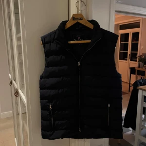 Svart dunväst från Bondelid  - Svart dunväst från Bondelid med horisontella quiltade paneler och hög krage. Västen har två fickor med dragkedja och hel dragkedja framtill. Insidan är slät och fodrad, perfekt för lager-på-lager under kyliga dagar men också som en helt vanlig väst. Knappt använd.