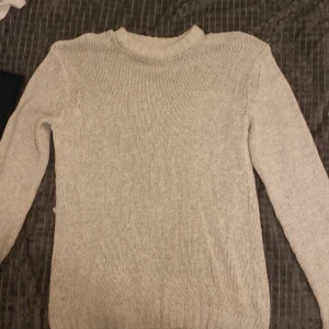 Beige stickad tröja med rund hals - En enkel och stilren beige stickad tröja med rund halsringning och långa ärmar. Tröjan har ribbade muddar vid ärmslut och nederkant, vilket ger en snygg och klassisk look. Perfekt att slänga på sig när det är kyligt ute.