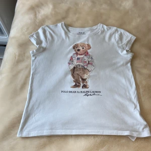 Vit Polo Bear t-shirt från Ralph Lauren - Vit t-shirt från Polo Ralph Lauren med ikoniskt Polo Bear-tryck framtill. Klassisk rund hals och korta ärmar. Tillverkad i mjuk bomull som är skön mot huden. Perfekt för dig som gillar stilrena plagg med en lekfull twist.