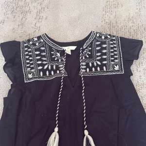 Svart blus med broderi från H&M - Svart blus från H&M med vita broderier och snygga detaljer vid axlarna. Blusen har korta volangärmar, v-ringning och dekorativa snören med tofsar framtill. Blusen säljs inte längre❤️