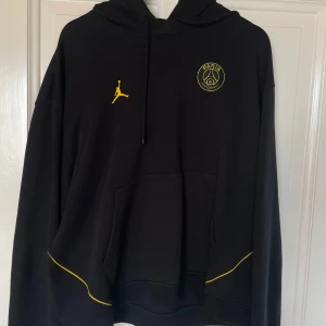 Svart PSG x Jordan hoodie XL - Säljer en svart hoodie från Jordan x Paris Saint-Germain med gula detaljer och stor logga på ryggen. Hoodien har huva, känguruficka och coola gula linjer längs sidorna. Perfekt för dig som gillar streetwear och fotbollsvibbar.