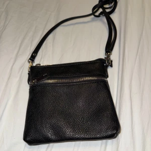 Svart axelväska med dragkedja - En stilren svart axelväska med justerbar rem och silverfärgade dragkedjor. Väskan har ett huvudfack och en ytterficka med dragkedja. Insidan är fodrad i svart tyg och har en extra innerficka med dragkedja.