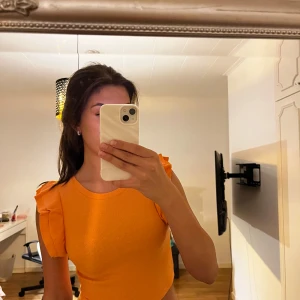 Croppad orange topp med volangärm - Säljer en croppad topp i stark orange färg med rund halsringning och korta ärmar med volangdetaljer. Materialet är ribbad och stretchigt, vilket gör den bekväm och snygg. Perfekt för dig som gillar färg och vill sticka ut.