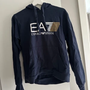 Mörkblå EA7 Emporio Armani hoodie - Snygg mörkblå hoodie från EA7 Emporio Armani med stor logga i vitt och guld på bröstet. Tröjan har huva med snörning. Bra skick. Anledning till försäljning är att den är för liten för mig 