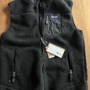 Svart fleeceväst från Patagonia - Svart fleeceväst från Patagonia med hög krage och hel dragkedja framtill. Västen har två sidofickor med dragkedja och en bröstficka med dragkedja och tygpatch med logga. Mjuk och fluffig känsla, perfekt för lager-på-lager-stil.