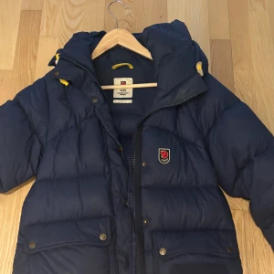 Blå dunjacka från Fjällräven - Säljer en blå dunjacka från Fjällräven med huva och gula detaljer. Jackan har dragkedja, tryckknappar och två stora fickor framtill. Klassisk Fjällräven-logga på bröstet och quiltad design som håller dig varm under kalla dagar.