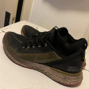 Nike Odyssey React Shield sneakers - Nike Odyssey React Shield sneakers i svart och olivgrön med vattenavvisande material och reflekterande detaljer. Skorna har snörning, robust sula med React-dämpning och röda detaljer undertill. Perfekta för löpning i alla väder.