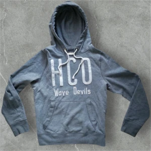 Unik Hollister Hoodie! - Säljer en grå hoodie från Hollister med stor vit HCO Wave Devils-tryck på bröstet. Tröjan har huva med vita snören och en stor magficka framtill. Perfekt för dig som gillar en avslappnad och sportig stil.