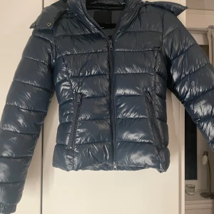 Mörkblå pufferjacka med huva - Säljer en snygg mörkblå pufferjacka med huva. Jackan har en glansig yta och dragkedja