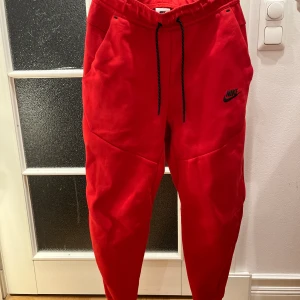 Röda byxor från Nike - Snygga röda mjukisbyxor från Nike med svart snörning i midjan. Byxorna har en avslappnad passform och är perfekta för en bekväm stil. Nike-loggan är tryckt på ena benet.