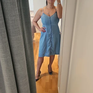 Denim klänning med knappar - Säljer en helt ny och söt ljusblå denimklänning med smala axelband och knappar framtill. Klänningen har en v-ringad design och är perfekt för varma dagar. Tillverkad i ett mjukt och bekvämt material som ger en skön passform. Passar bra för sommarens alla tillfällen. Strecth vid ryggen. Prislapp finns kvar. 