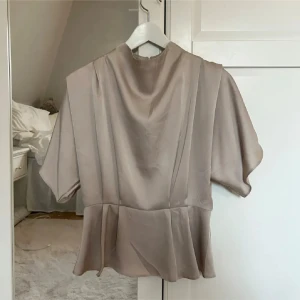 Beige blus med korta ärmar - Elegant beige blus med korta ärmar och en lös passform. Blusen har en mjuk och flytande design med en lätt drapering vid halsen. Perfekt för en stilren och sofistikerad look. Tröjan är i storlek L men passar även M.