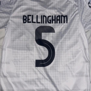 Vit Real Madrid fotbollströja - Säljer en vit Real Madrid fotbollströja från Adidas med korta ärmar. Tröjan har klubbens emblem och sponsorlogga på framsidan samt spelarnamnet Bellingham och nummer 5 på baksidan. Perfekt för fans av laget!