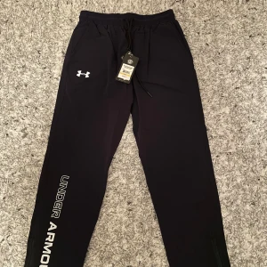Svarta mjukisbyxor från Under Armour - Snygga svarta mjukisbyxor från Under Armour med logga på benet. Byxorna har en elastisk midja med snörning för bästa passform. Perfekta för träning eller en avslappnad dag hemma. Passar mer som M-L