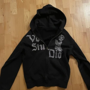 Vonstudio zip - Snygg svart hoodie med dragkedja och rhinestones som bildar text och en figur på framsidan. Perfekt för en cool och avslappnad stil. Den har en klassisk passform med långa ärmar och en praktisk huva.