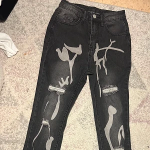 Svarta jeans med abstrakt mönster - Unika svarta jeans med abstrakt mönster i grått och slitna detaljer. Högmidjade med en straight passform. Perfekta för dig som vill sticka ut med en edgy stil.