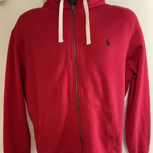 Röd hoodie från Ralph Lauren - Snygg röd hoodie från Ralph Lauren med dragkedja och vit snörning i huvan. Den har en liten blå logga på bröstet och praktiska fickor framtill. Perfekt för en avslappnad stil.