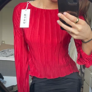 Röd plisserad blus från NA-KD med öppen rygg - Trendig röd blus från NA-KD med plisserad design och öppen rygg. Blusen har långa ärmar och knytband i ryggen för en elegant touch. Aldrig använt, lappen är kvar!