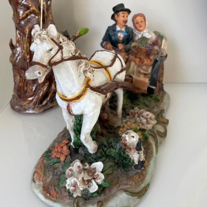 Dekorativ figur av häst och vagn - Charmig dekorativ figur föreställande en häst och vagn med ett par klädda i gammaldags kläder. Detaljerad med blommor och en hund vid basen. Perfekt som prydnad i hemmet..köpt år 1990.måttet längd är 38cm och bredden är 15cm .kan möta er i Stockholm utanför Åhléns i T-Centralen 