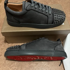 Christian Louboutin skor - Snygga grå sneakers från Christian Louboutin med nitar på tån och den ikoniska röda sulan. Skorna har snörning och är tillverkade i ett stilrent material som ger en lyxig känsla.