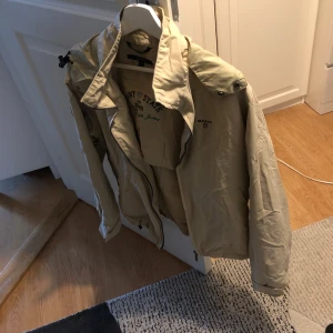 Beige jacka från Gant - Säljer en beige jacka från Gant med broderad text på insidan. Jackan har en klassisk design med dragkedja och knappar. Perfekt för vår och höst med sin stilrena look.
