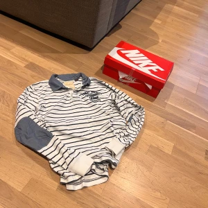 Randig pikétröja med krage - Säljer en stilren vit och blå randig pikétröja med lång ärm och kontrasterande krage. Tröjan har en broderad detalj på bröstet och är perfekt för en avslappnad look. Passar bra till jeans eller chinos.