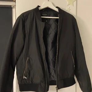 Svart bomberjacka lager 157 - Snygg svart bomberjacka från lager 157 med dragkedja framtill och ribbade muddar. Jackan har en quiltad insida och två fickor med dragkedja. Perfekt för en stilren look.