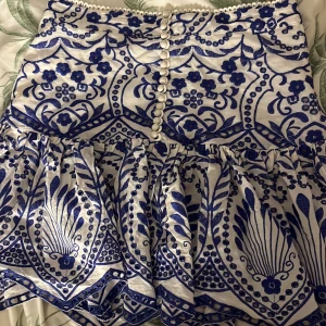 Vit och blå kjol med blommönster - Snygg vit och blå kjol med ett detaljerat blommönster och knappar framtill. Kjolen har en volangdetalj som ger extra stil och rörelse. Perfekt för att sticka ut med en unik look.