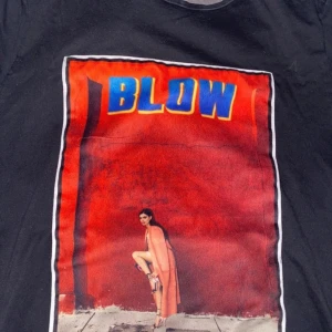 Svart t-shirt med tryck från Limitato - Snygg svart t-shirt från Limitato med ett färgstarkt tryck av en kvinna mot en röd bakgrund och texten 'BLOW' i blått och gult. Perfekt för dig som vill sticka ut med en unik design.