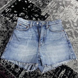 Blå jeansshorts från Weekday - Snygga blå jeansshorts från Weekday med fransiga kanter och klassisk femficksdesign. Perfekta för en avslappnad stil. De har en knappgylf och är tillverkade i ett slitstarkt denimtyg.